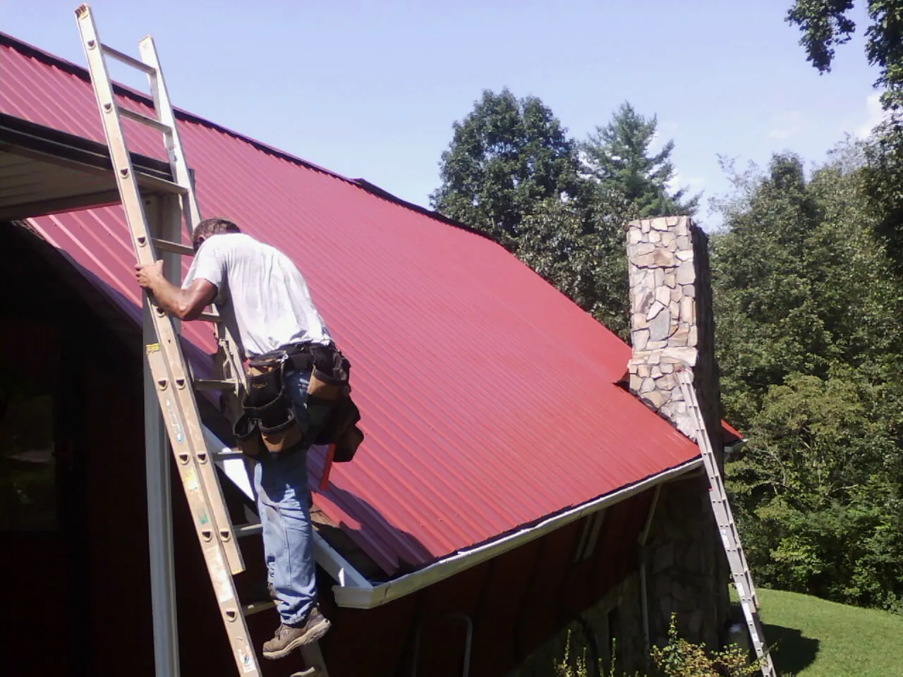 Roofer on a ladder beside a red metal roof and stone chimney — Soffit & Fascia Repair by Roofing Brothers in Oregon